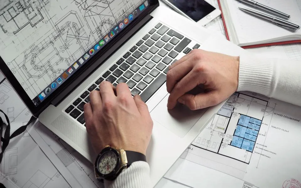 Hands working at a laptop showing a technical drawing, resting on printed plans