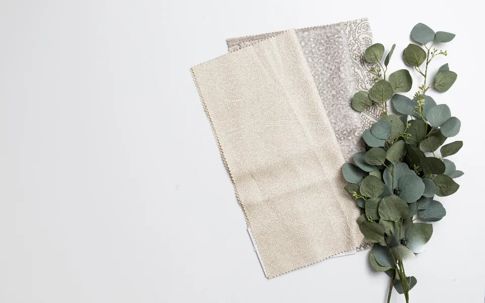 A plain cream fabric swatch overlapping a patterned grey one on a white surface, beside a eucalyptus sprig