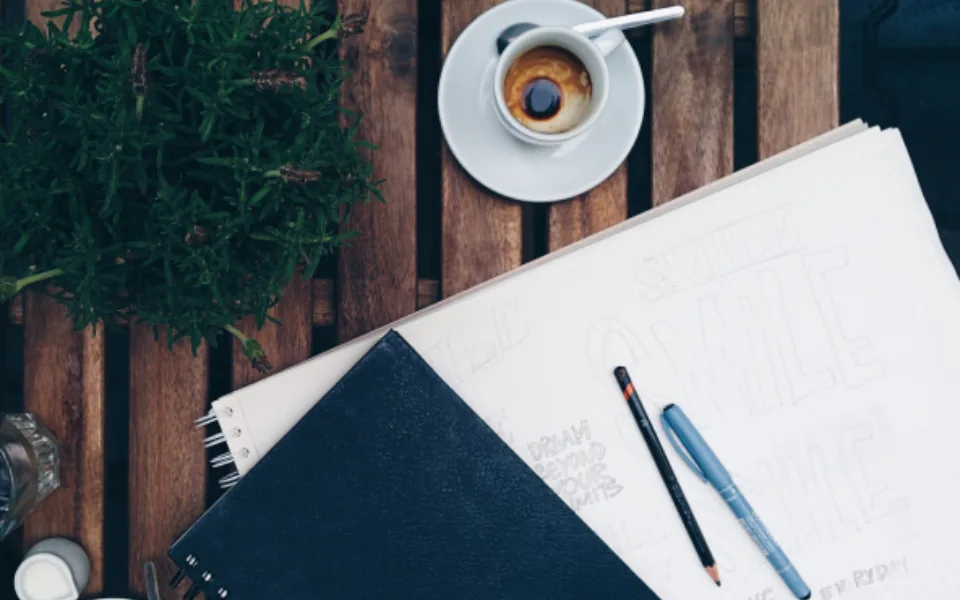 A sketchpad of hand-lettering under a closed notebook on a wooden table, with a pencil, a pen and an espresso