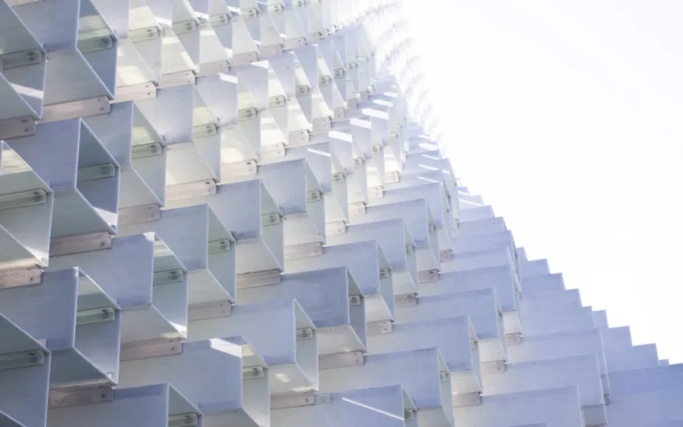 A pale facade of folded metal fins in a repeating grid, daylight showing in the gaps between them